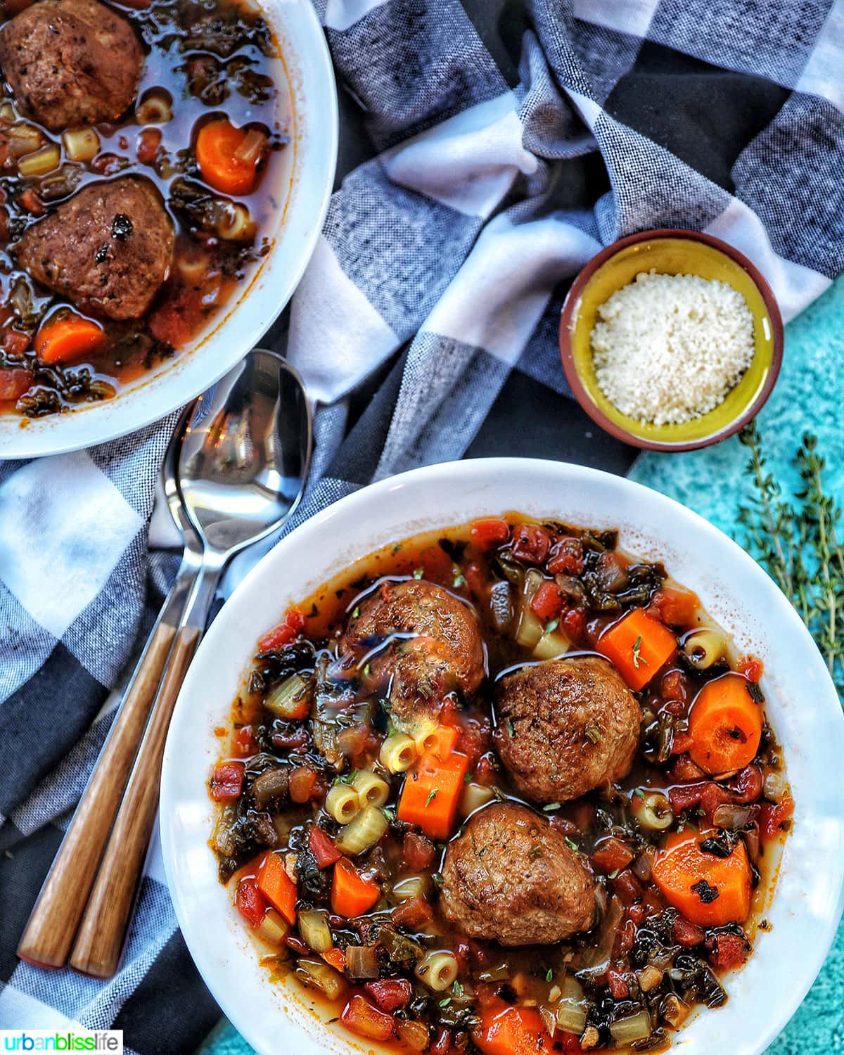 large white bowl of meatball soup with carrots, pasta, and a side of parmesan cheese and spoons.