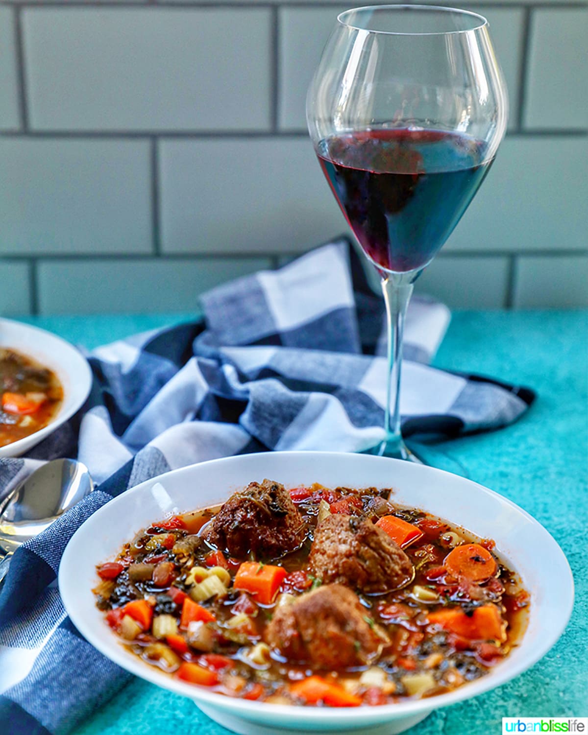 glass of red wine with bowl of meatball soup on a blue table with checkered napkin.