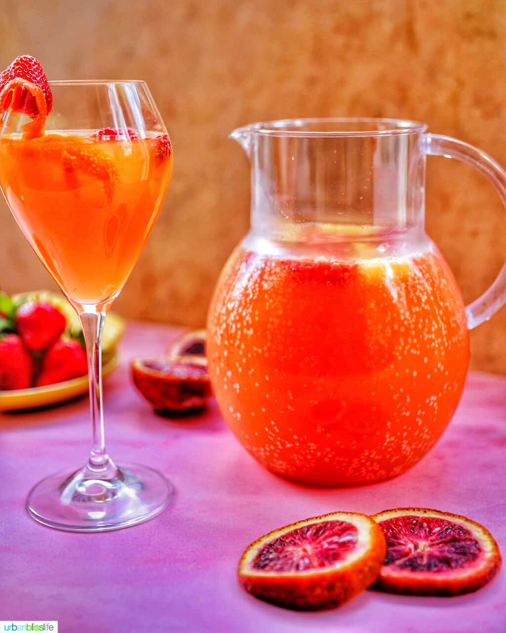 glass of tropical sangria with raspberries and citrus on a pink table with berries and a pitcher of sangria in the background.