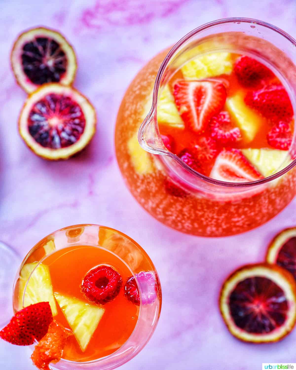 pitcher and glass of tropical sangria with strawberries, pineapples and raspberries floating in the drinks, on a pink table, with blood orange slices in the background.