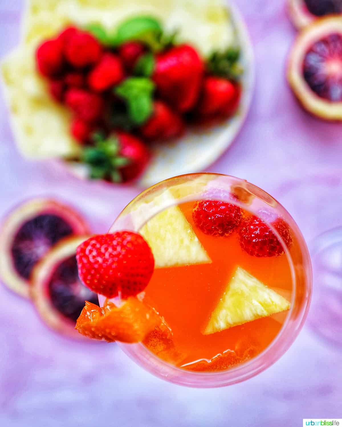 glass of tropical sangria with fruit slices in the drink on a pink background with fruit on a plate in the background.