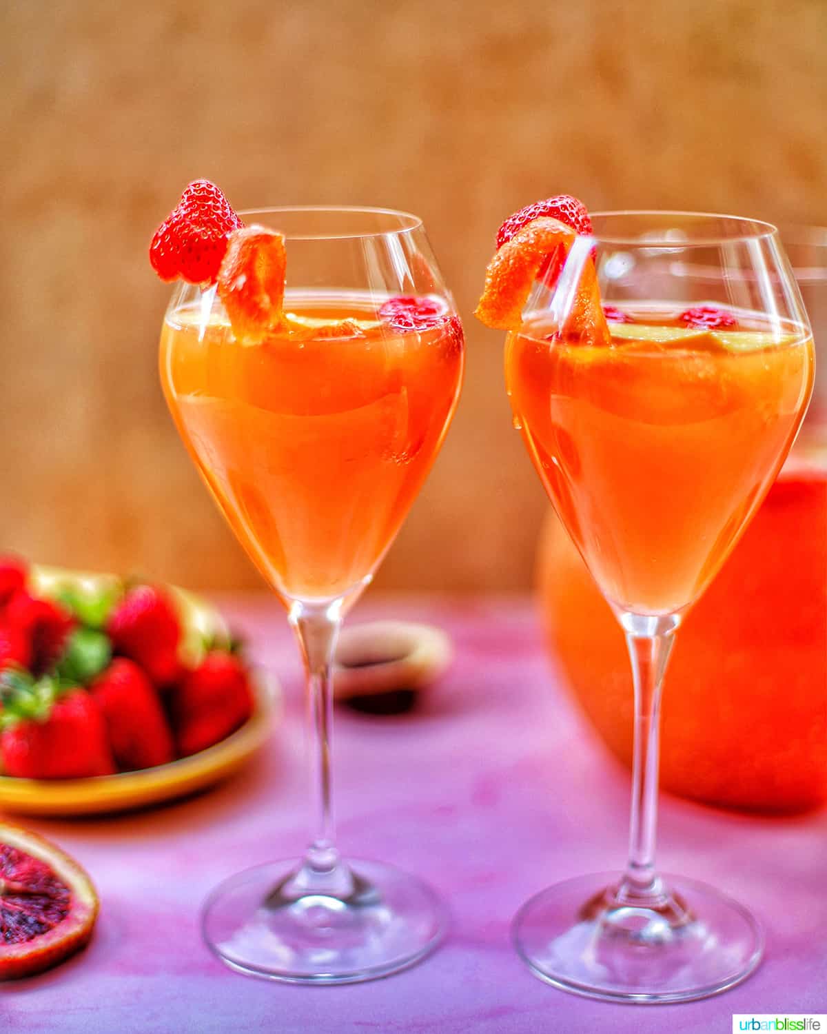 two glasses of tropical sangria with raspberries and citrus on a pink table with berries and a pitcher of sangria in the background.