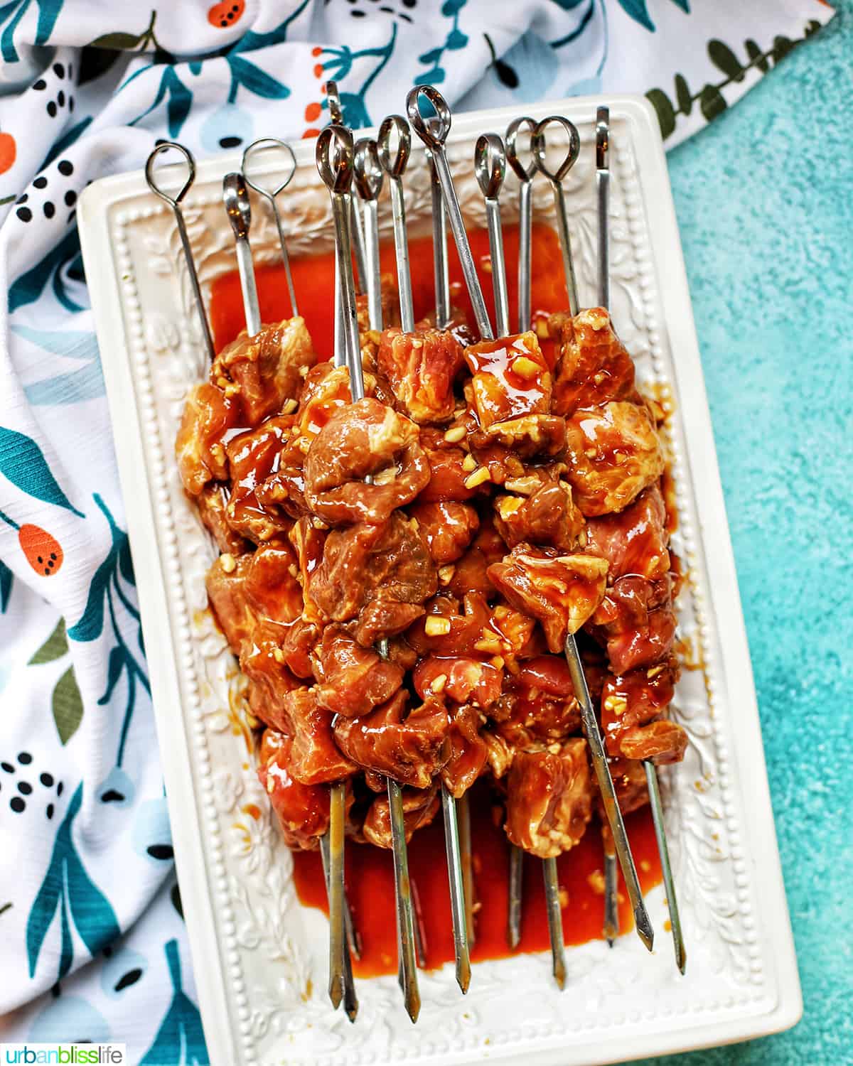 raw Filipino BBQ pork skewers on a white platter with a colorful napkin, all on a bright blue background.