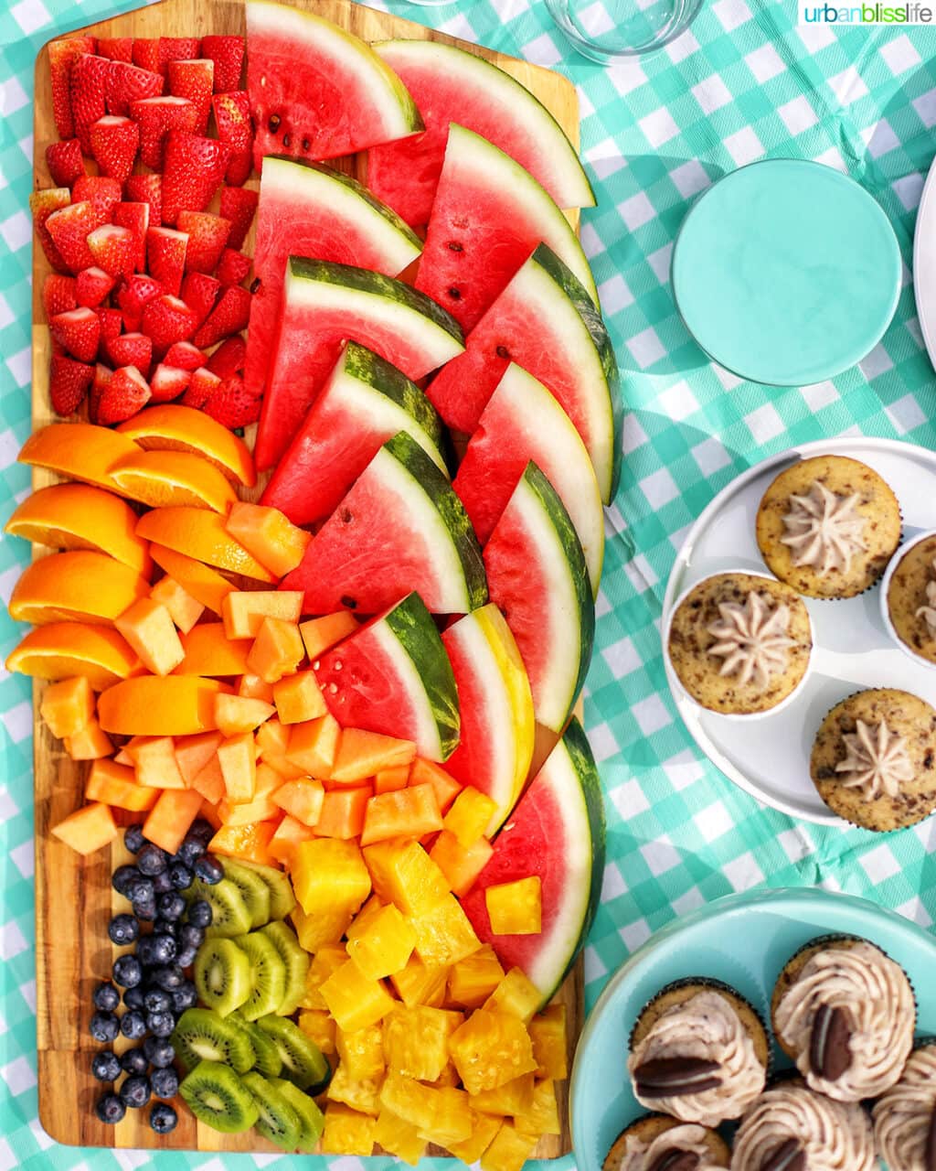 large fruit platter on an aqua checkered tablecloth with side of cupcakes