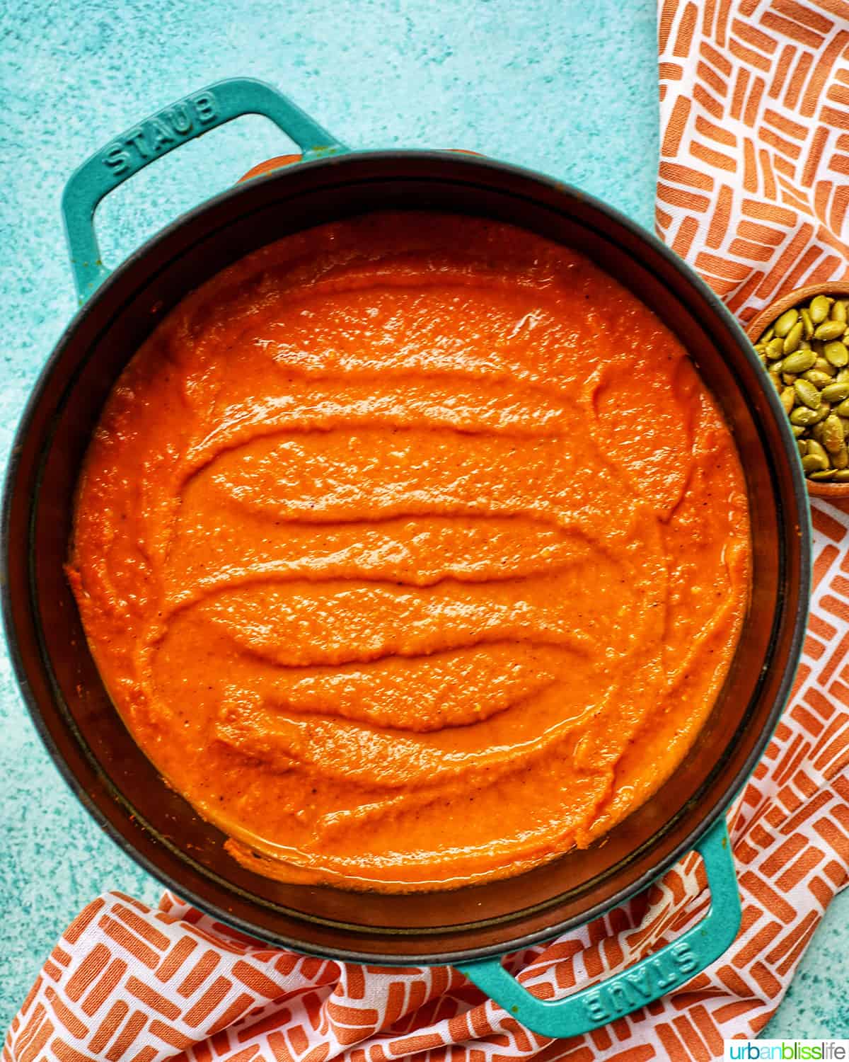 bright orange vegan pumpkin soup in staub pot on a blue background with side of pumpkin seeds.