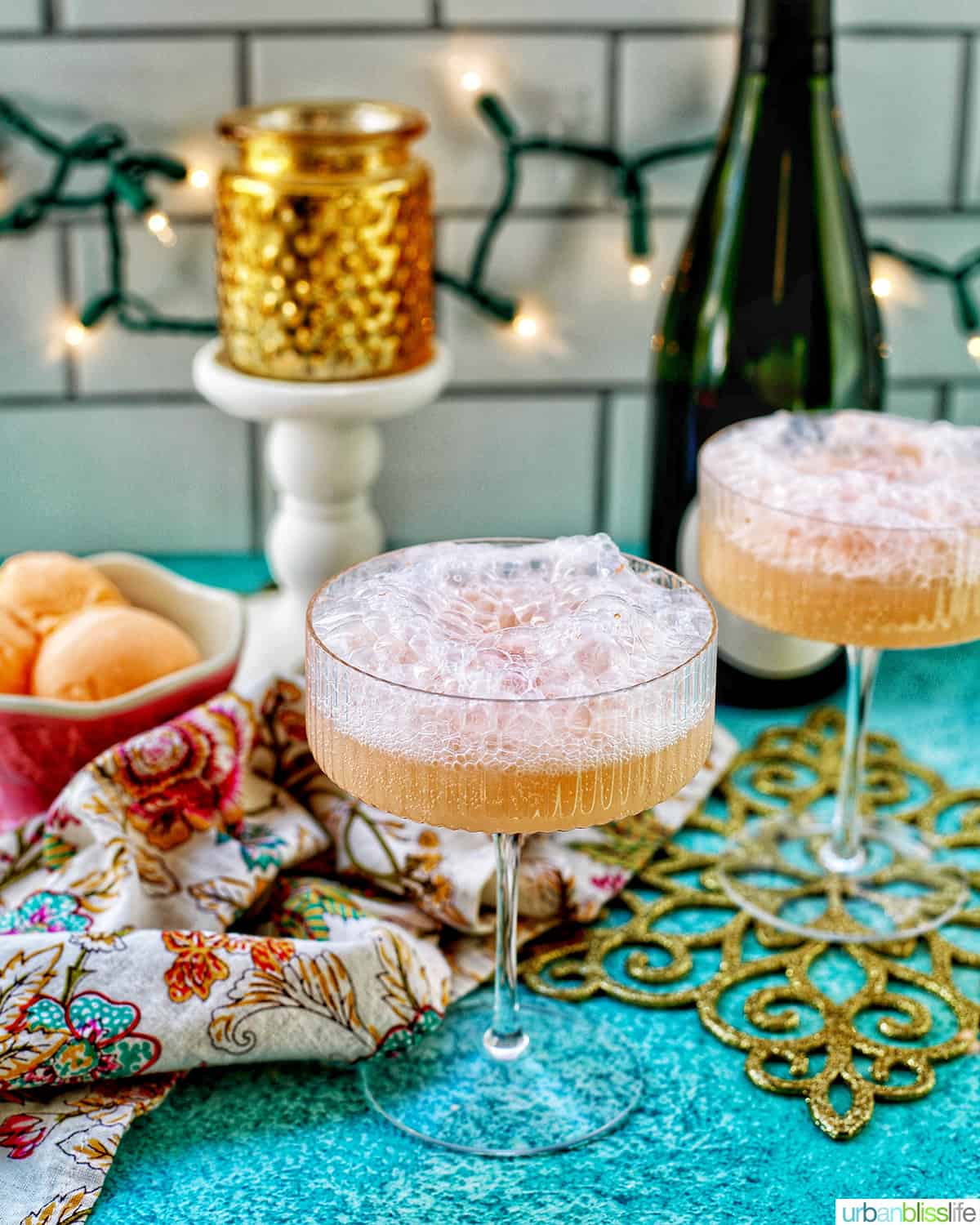 two glasses of champagne sherbet floats with twinkling lights and Champagne bottle in background and colorful decor.
