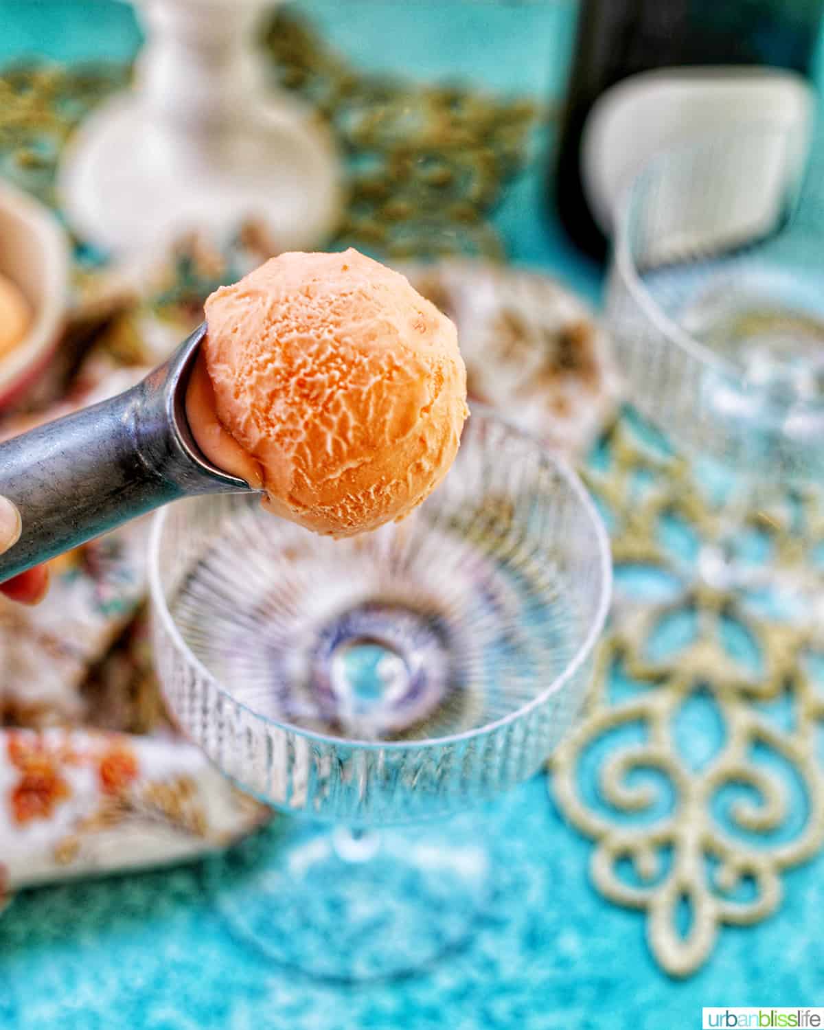 scooping orange sherbet into champagne class with gold decor and blue table blurred in the background.