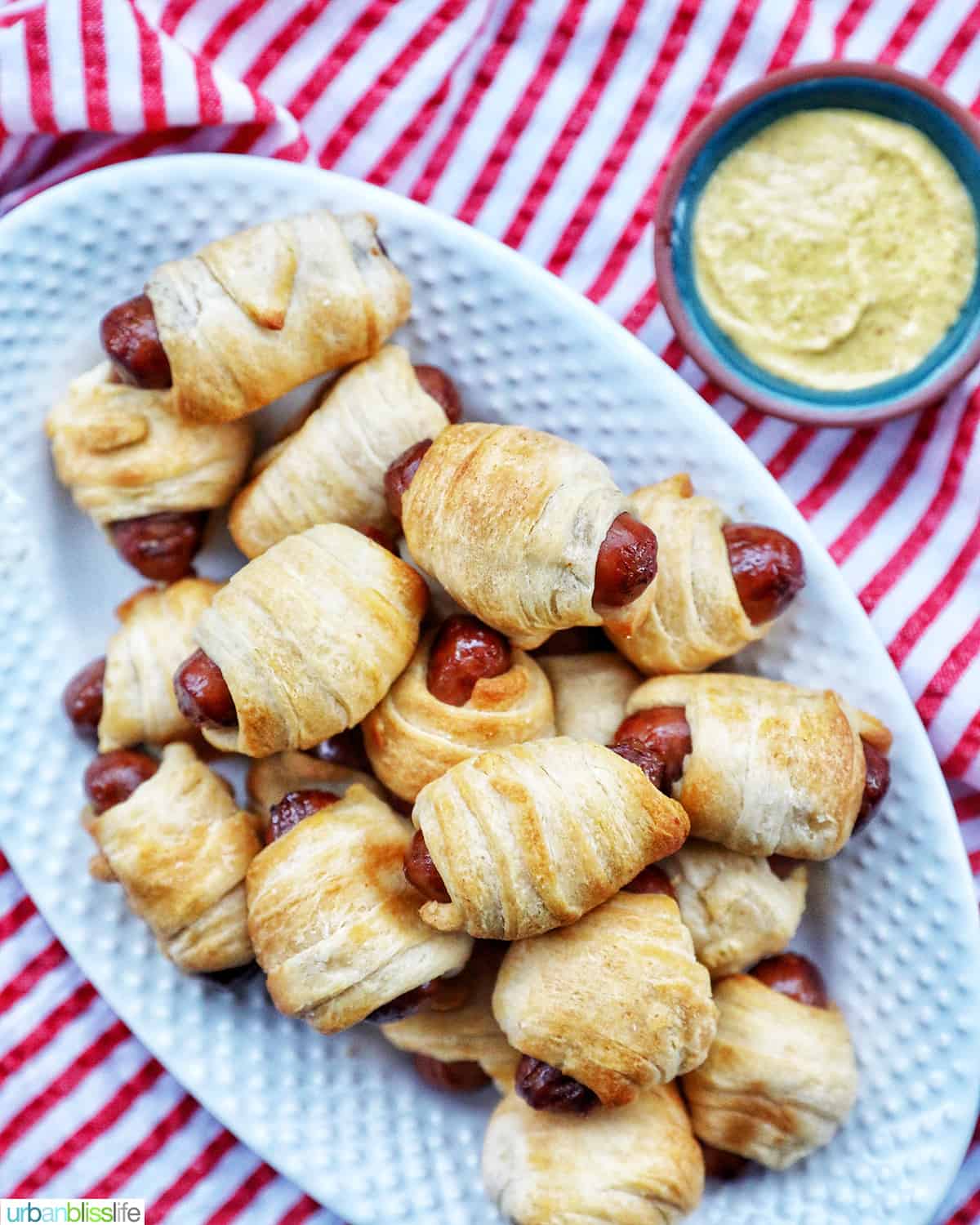 dozens of Air Fryer Pigs in a Blanket on a plate with side of mustard on a red and white napkin.