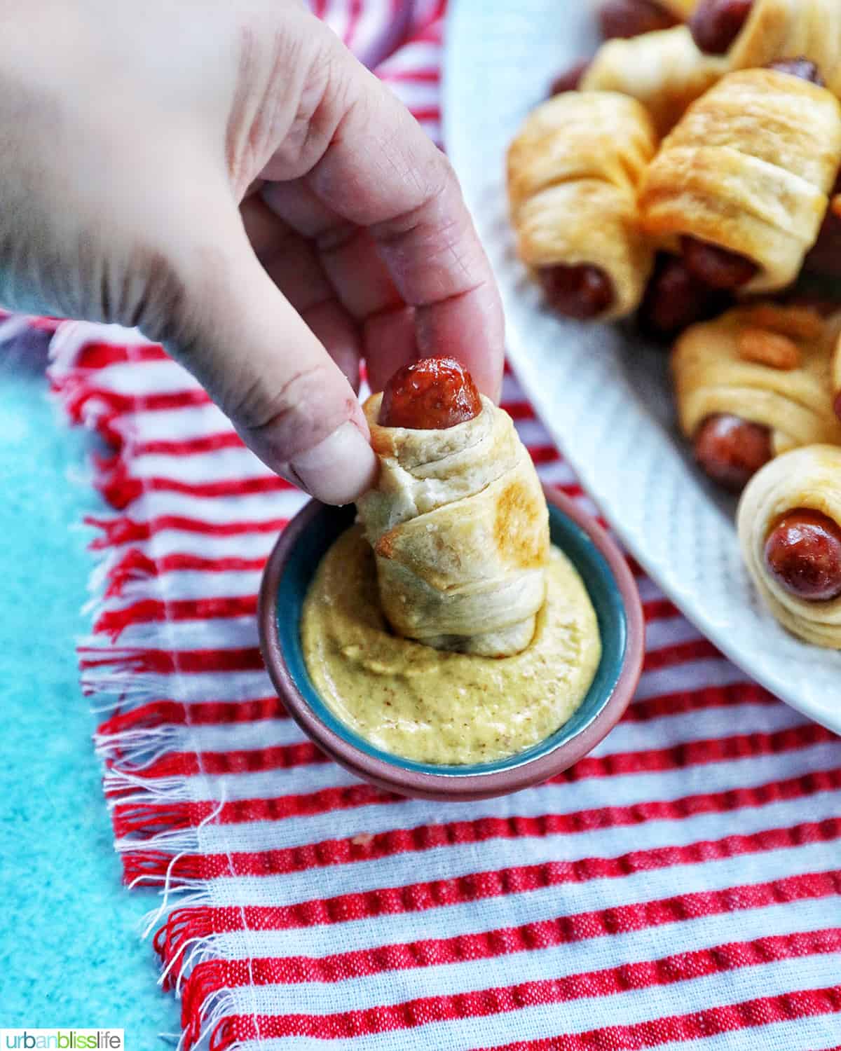 a hand dipping Air Fryer Pigs in a Blanket into mustard on a red and white napkin with several pigs-in-a-blanket on a plate.