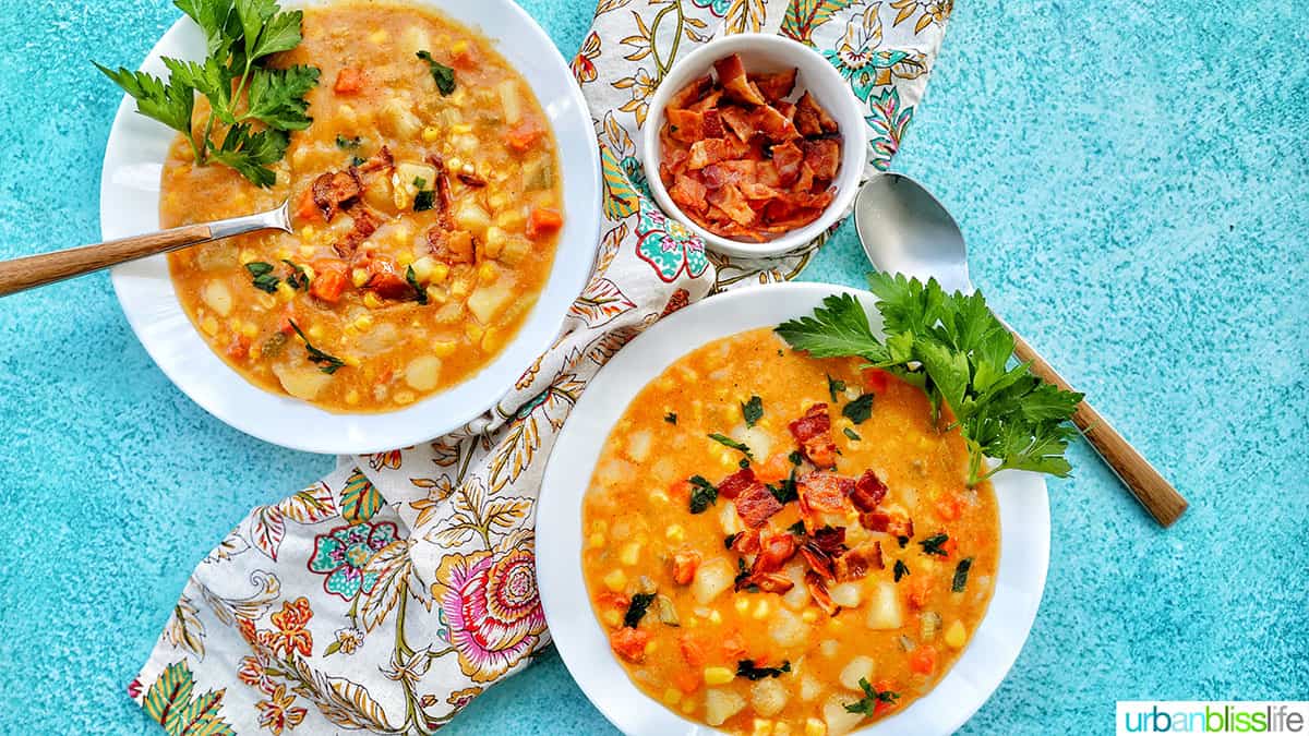 two bowls of Instant Pot Potato Corn Chowder with chopped bacon and side of parsley & bacon crumbles, and two spoons on a bright blue background with colorful floral napkin.