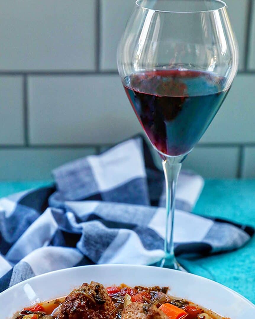 glass of red wine with bowl of soup and white and blue striped napkin.