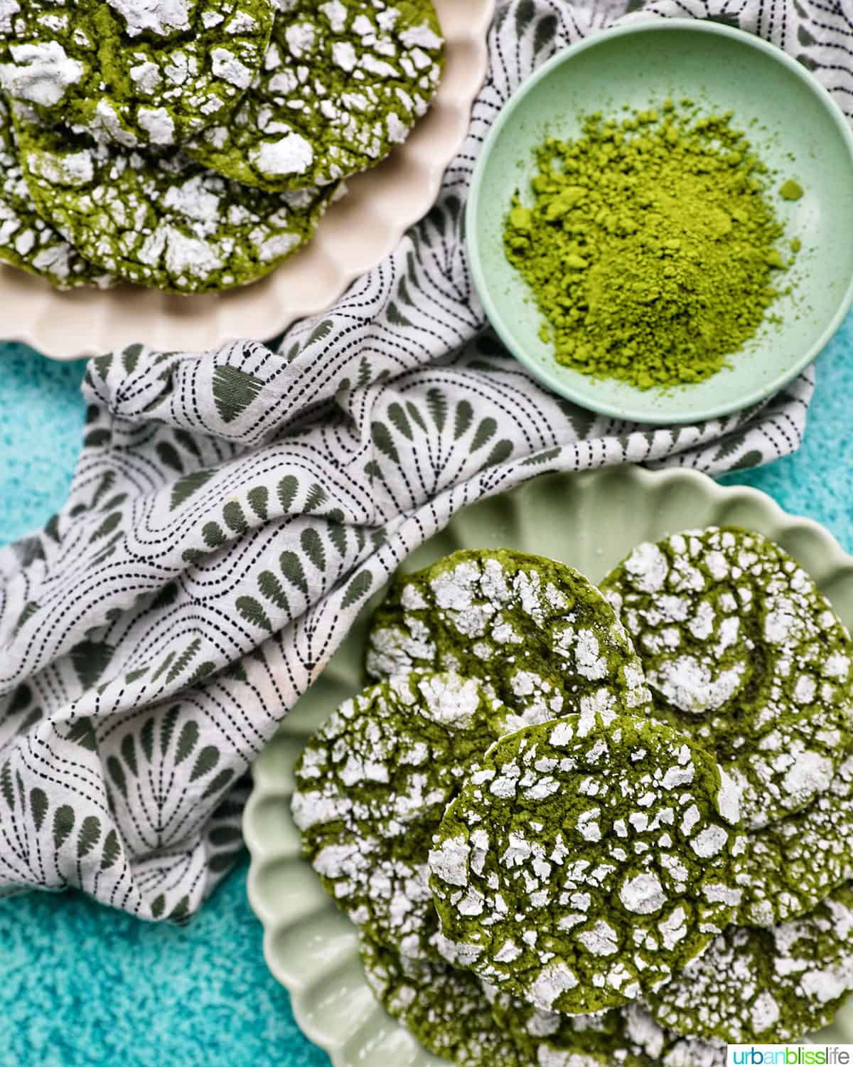 closeup flatlay of two plates of bright green matcha crinkle cookies with smaller plate of matcha powder.