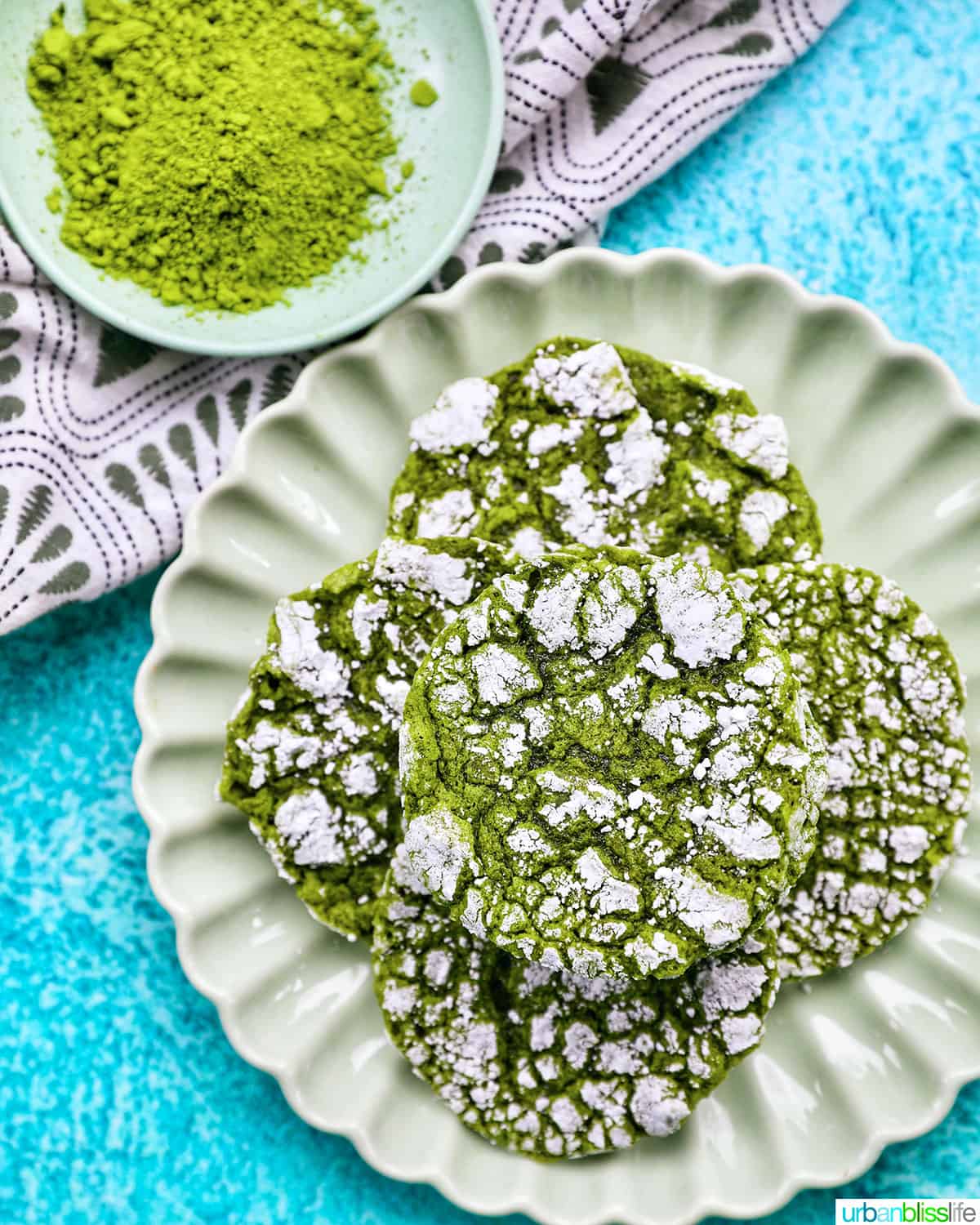 plate of bright green matcha crinkle cookies stacked on top of one another on a bright blue background.