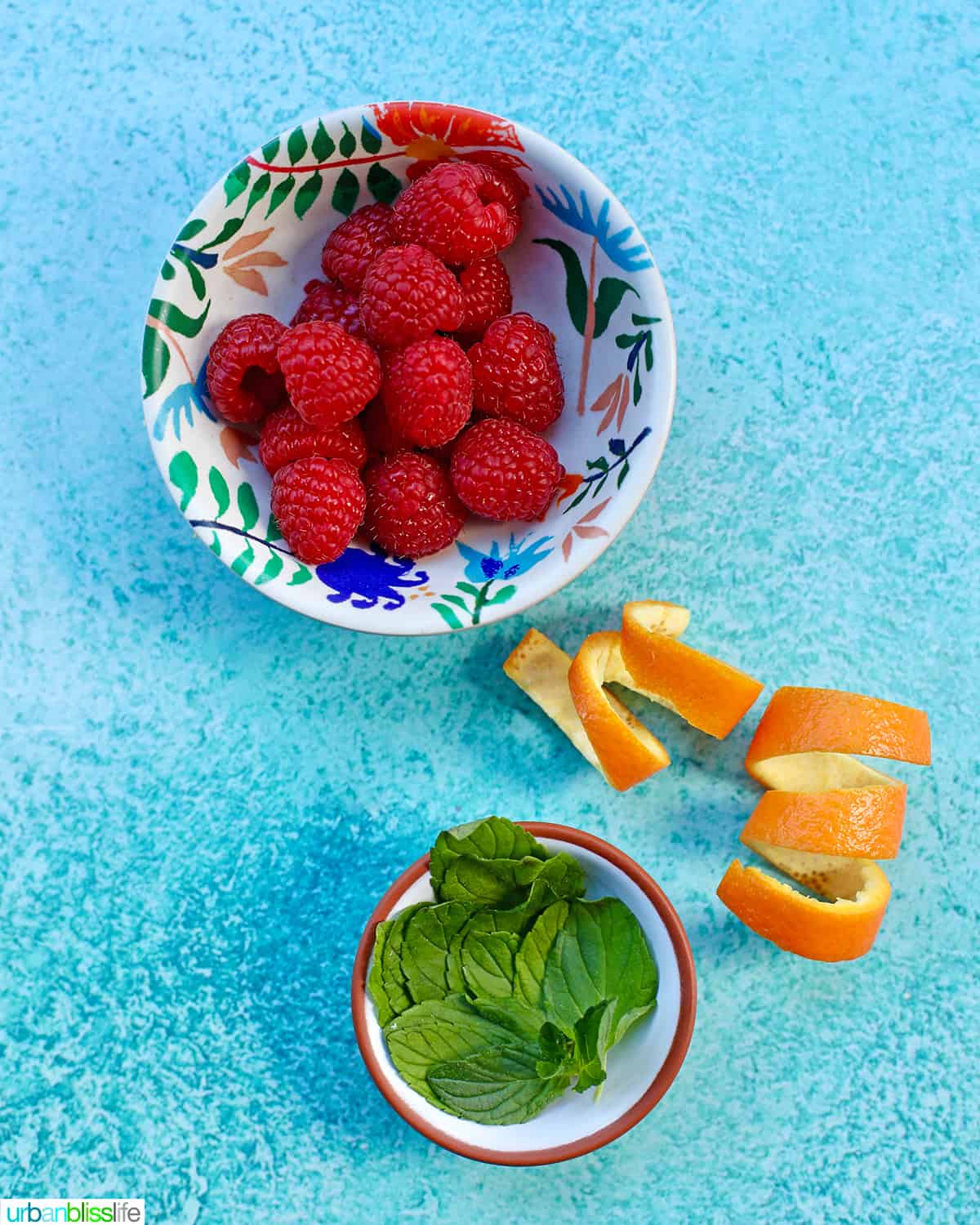 bowls of raspberries, mint leaves, orange rind garnishes for raspberry mimosas on blue background.