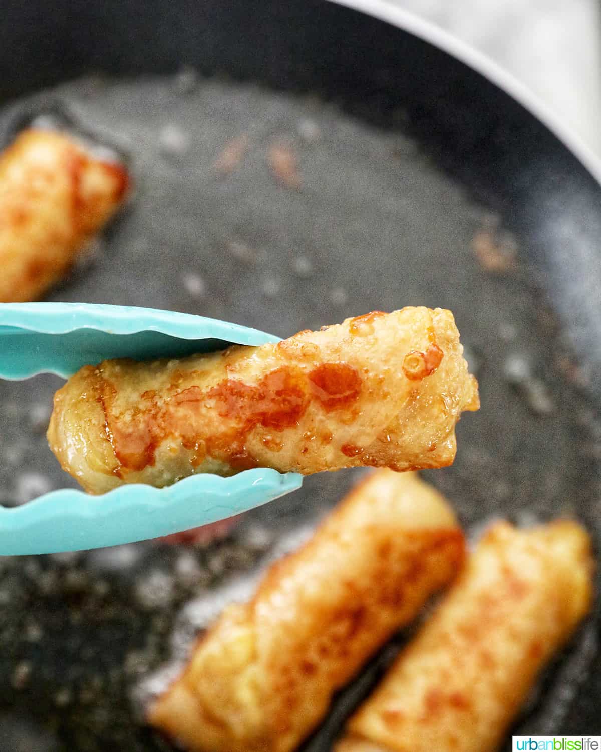 tong holding up a fried banana turon lumpia with frying more Filipino turon frying in the background.