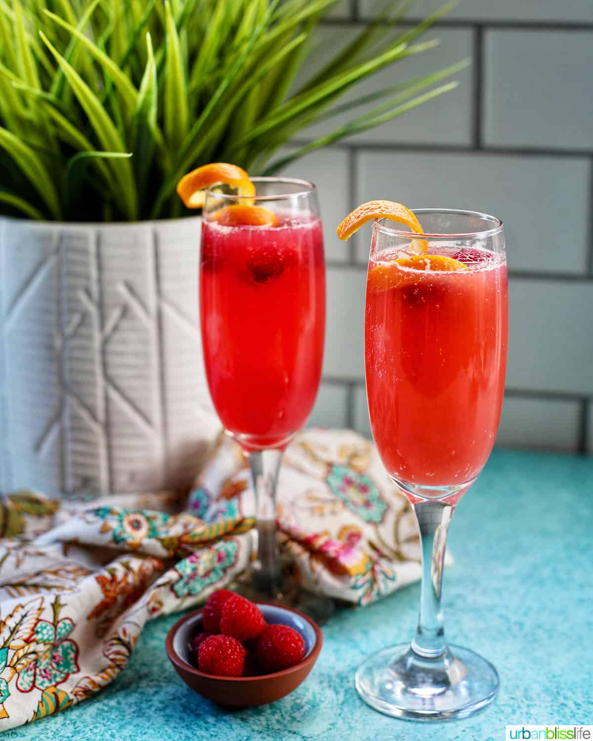 two glasses of Raspberry Mimosas Raspberry Mimosa with raspberries and orange rind garnish on blue background.