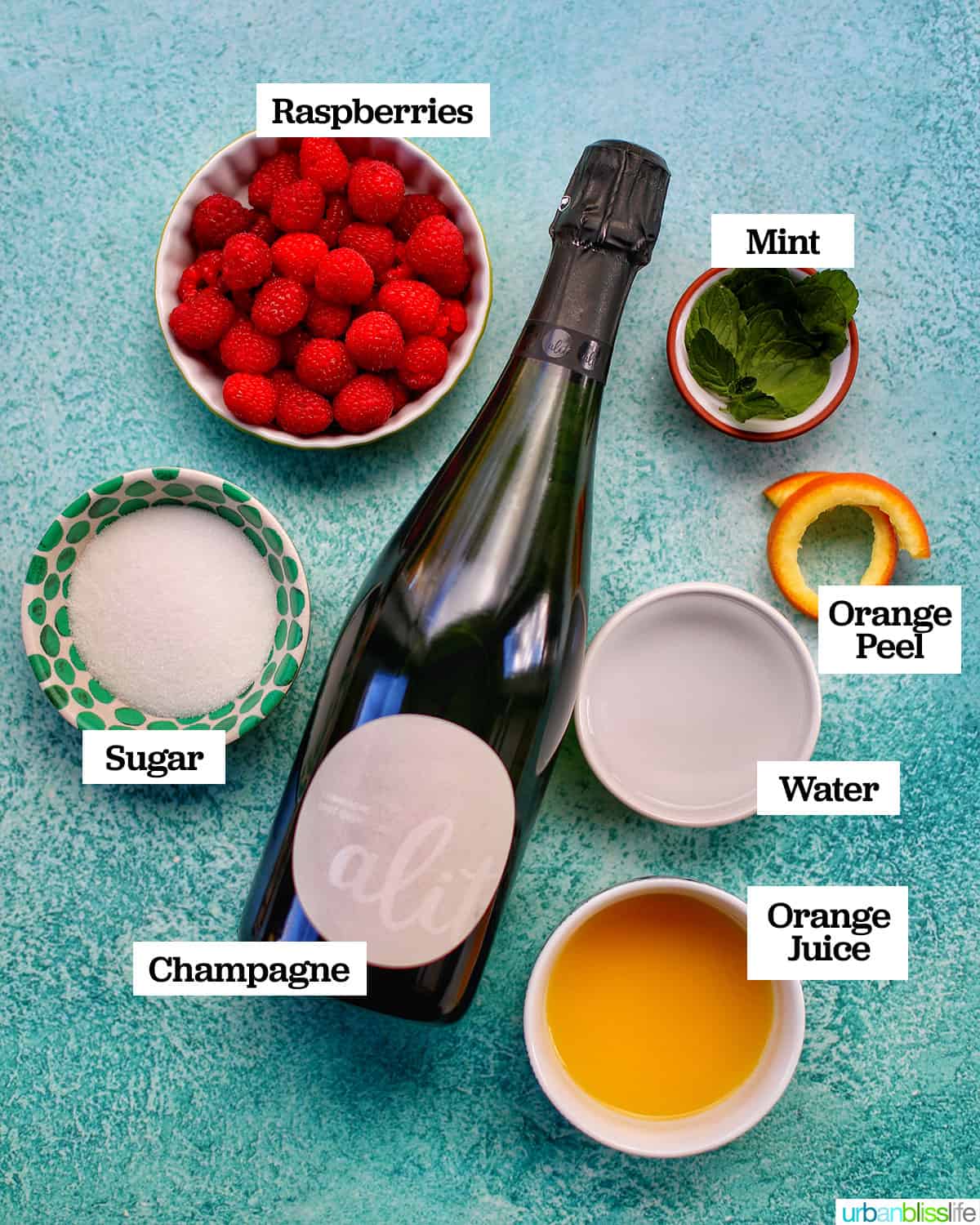ingredients needed to make Raspberry Mimosas in bowls with glass of sparkling wine on blue table.