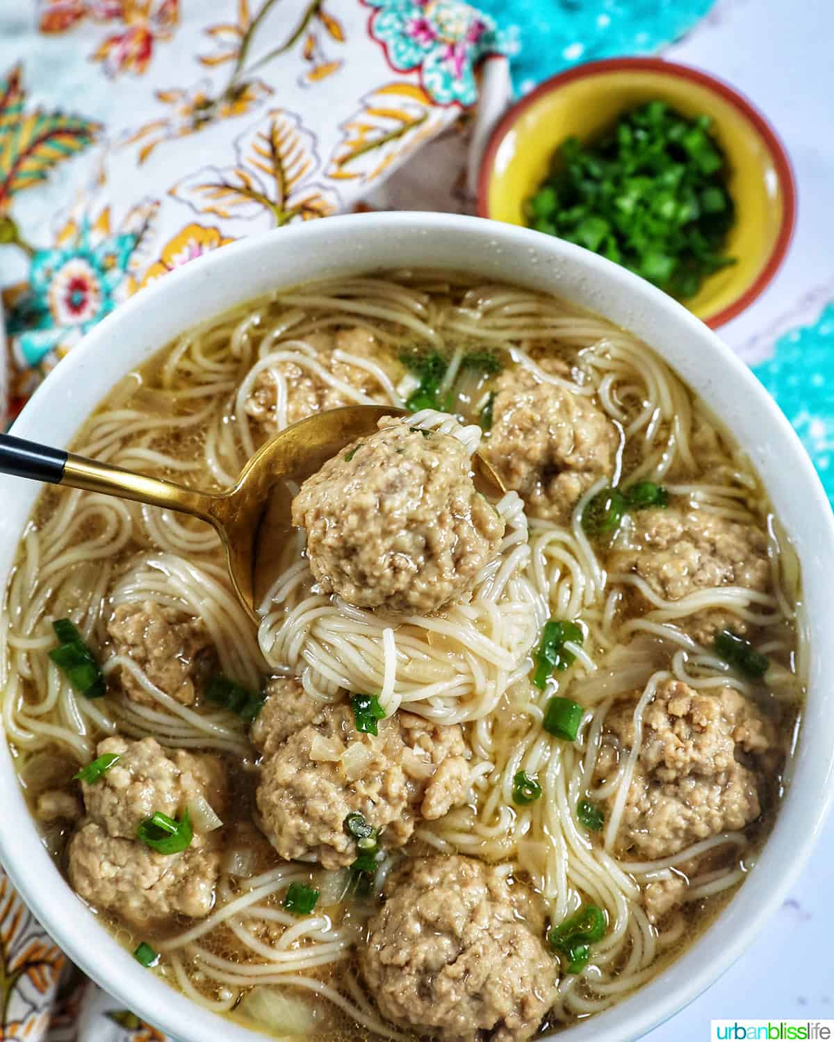 spoonful of filipino almondigas lifting out of a white bowl of meatball and noodle soup, with chopped green onions. 