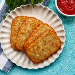 hash brown patties on a white plate with side of ketchup, greens, and blue background.