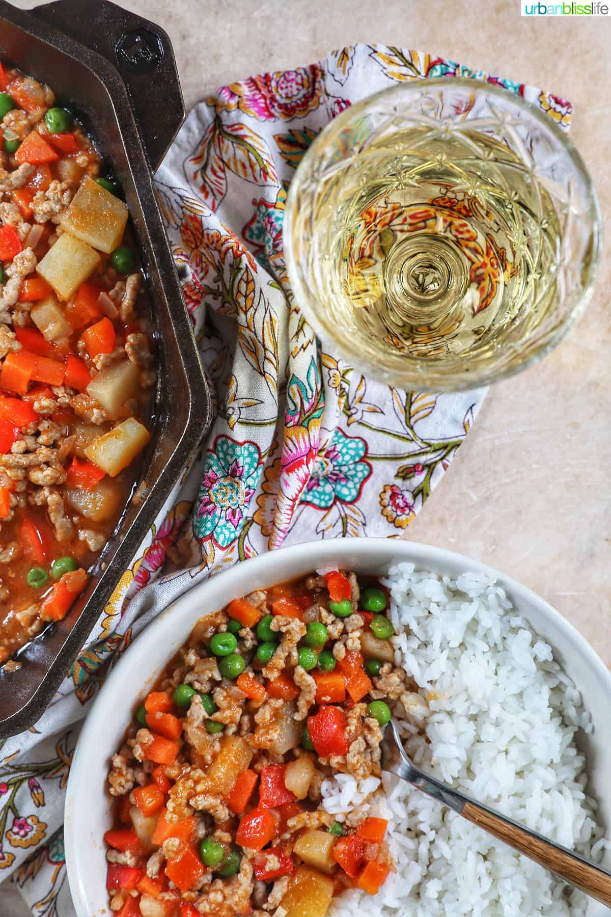 glass of white wine with bowls of Filipino menudo on a table with floral napkin.
