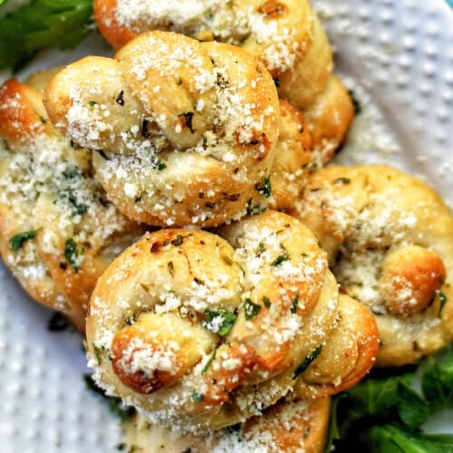 garlic bread rolls with parmesan cheese and parsley stacked on top of a white plate and bed of greens.