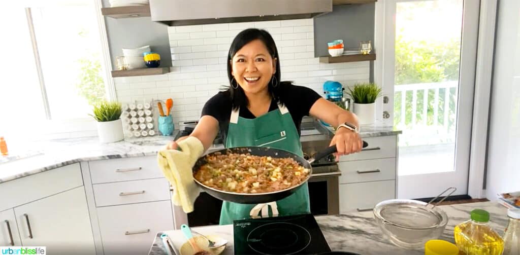 Marlynn Jayme Schotland holding a pan of lumpia filling in a white kitchen.