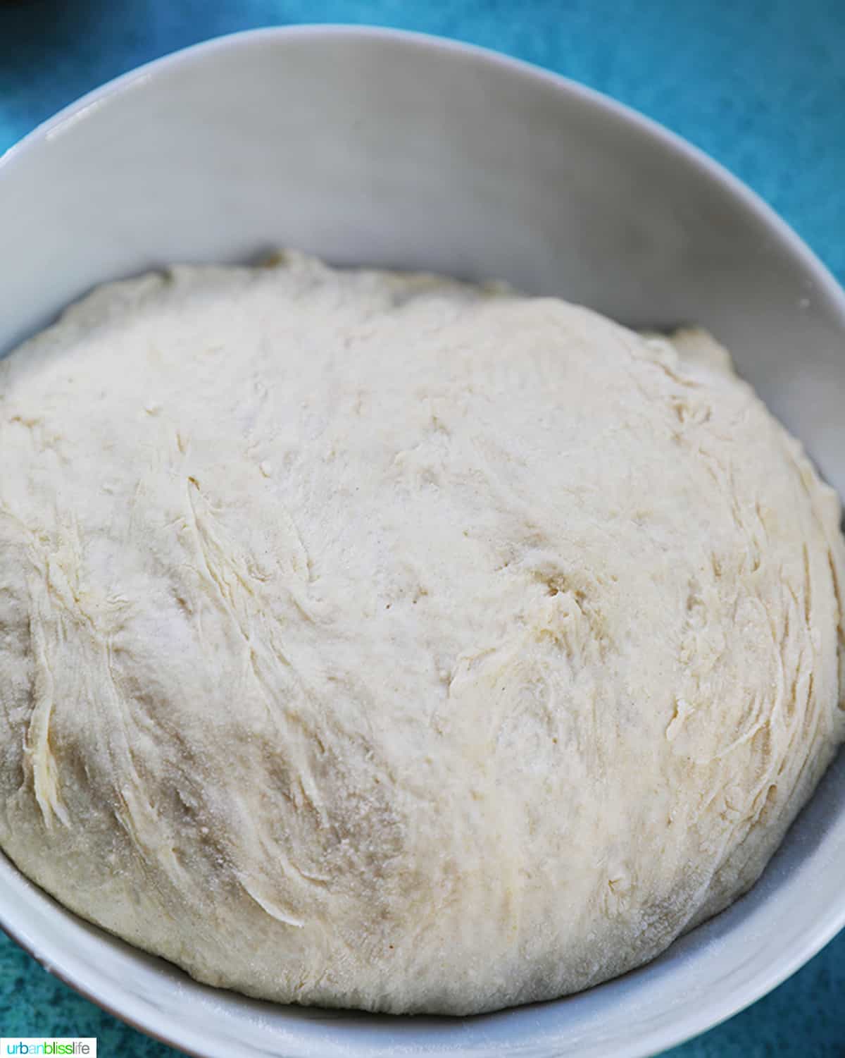 proofed pizza dough, raw, in a white bowl on blue table.