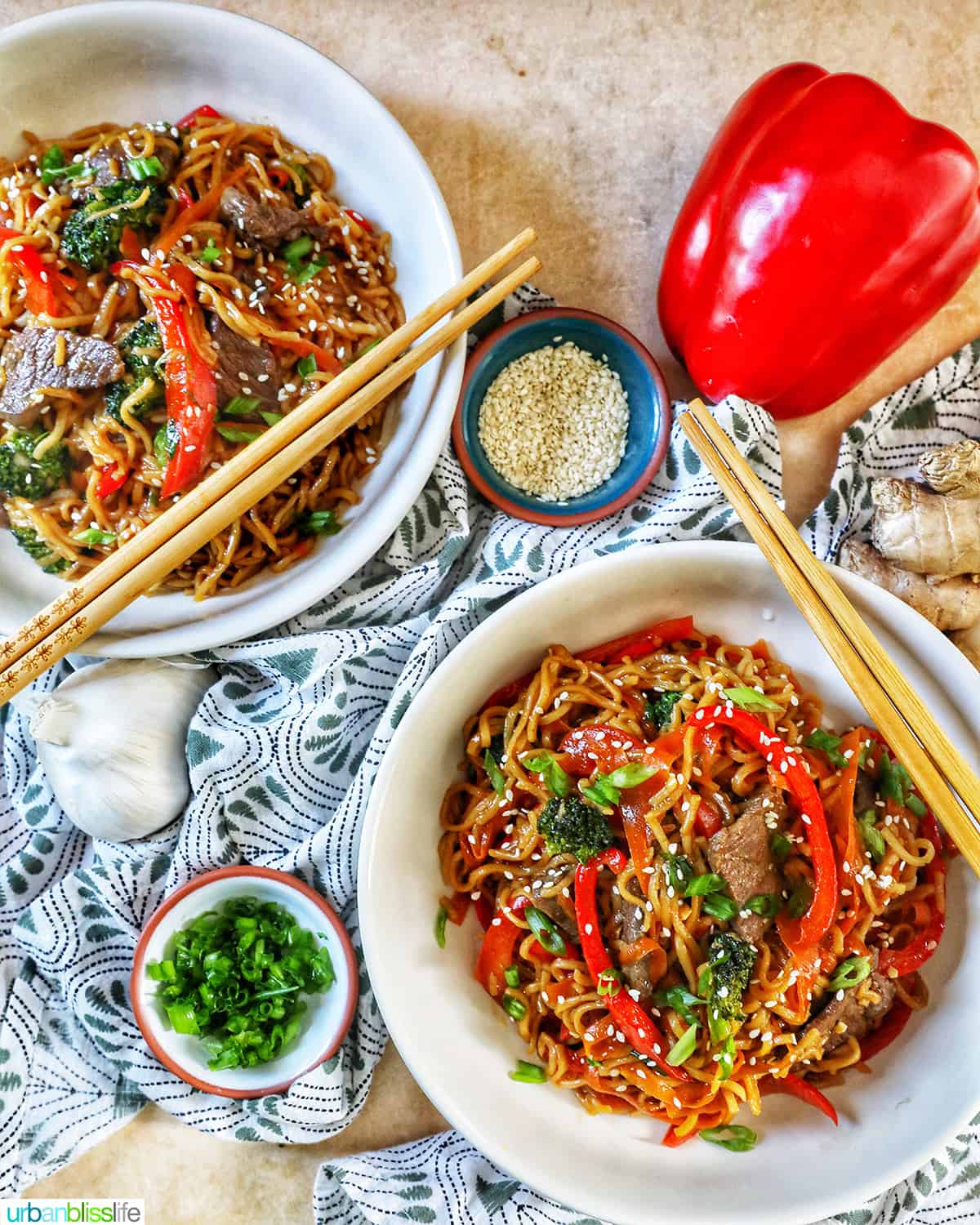 two bowls of sesame garlic noodles with chopsticks, bowls of scallions and sesame seeds, a red bell pepper, all on a green and white napkin and yellow table.