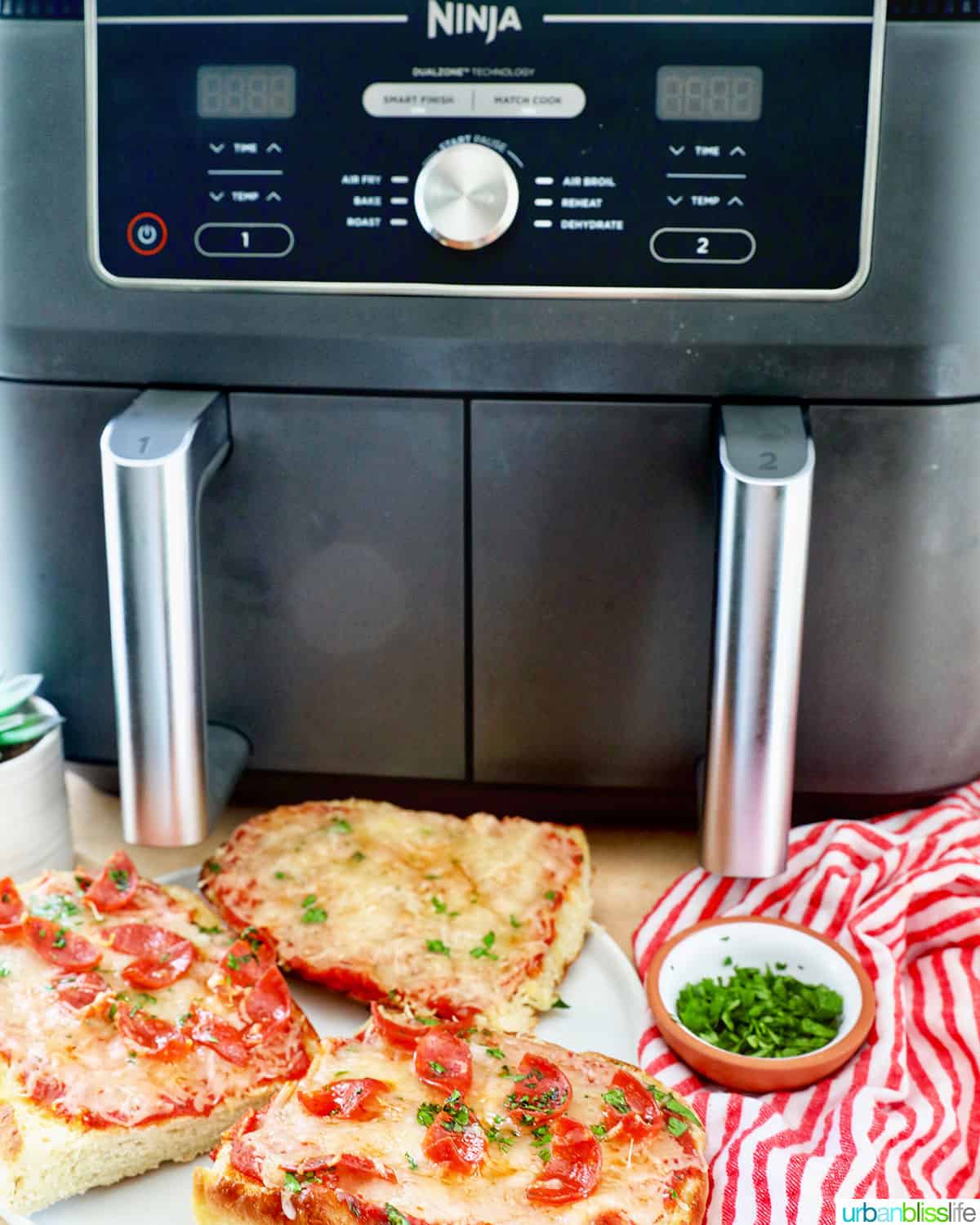 Ninja Foodi dual air fryer with french bread pizza and bowl of parsley in front on a red striped napkin.