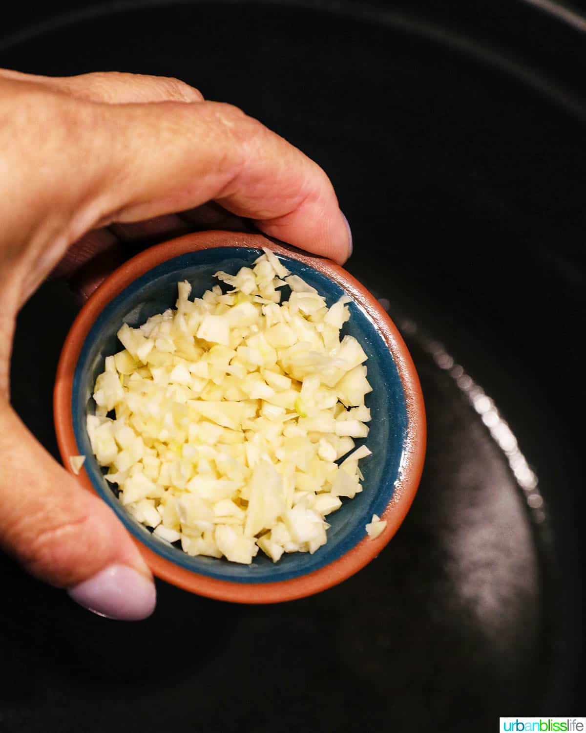 hand holding a pinch bowl full of minced garlic adding to a dutch oven pan or pot.