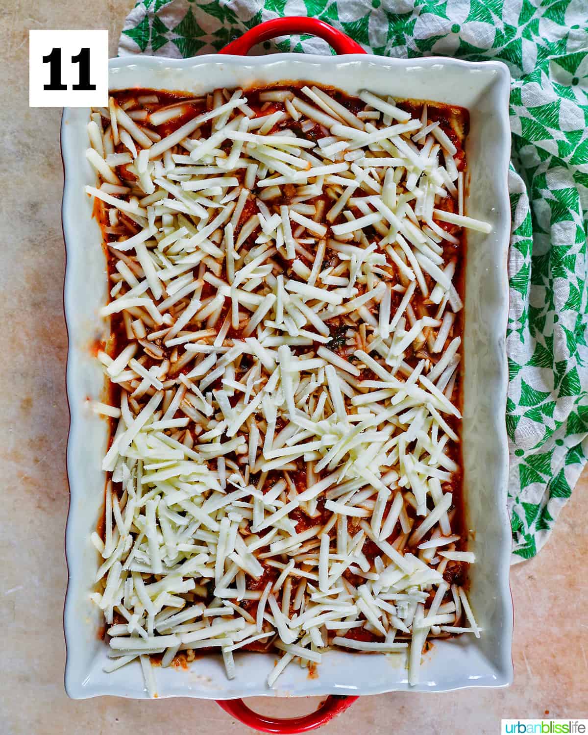 shredded dairy free mozzarella cheese added to the top layer of dairy free lasagna in a baking dish with red handles.