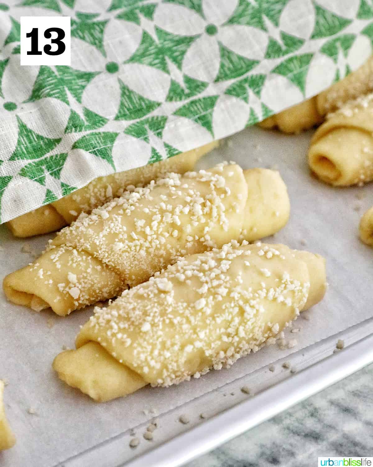 rolled spanish bread on a baking sheet covered with green and white napkin for a second rise.