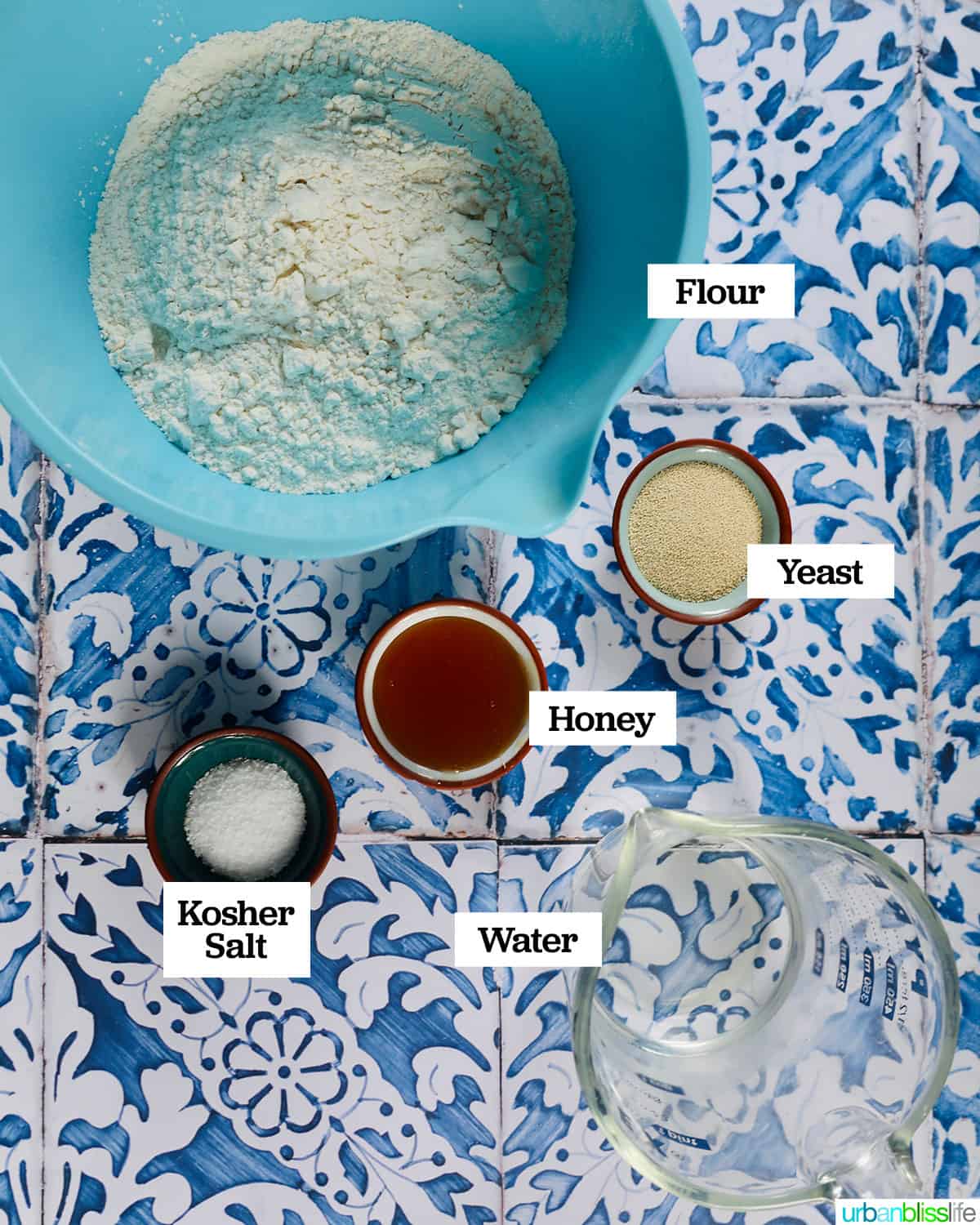 bowls of ingredients to make homemade french bread on a blue and white tiled table.