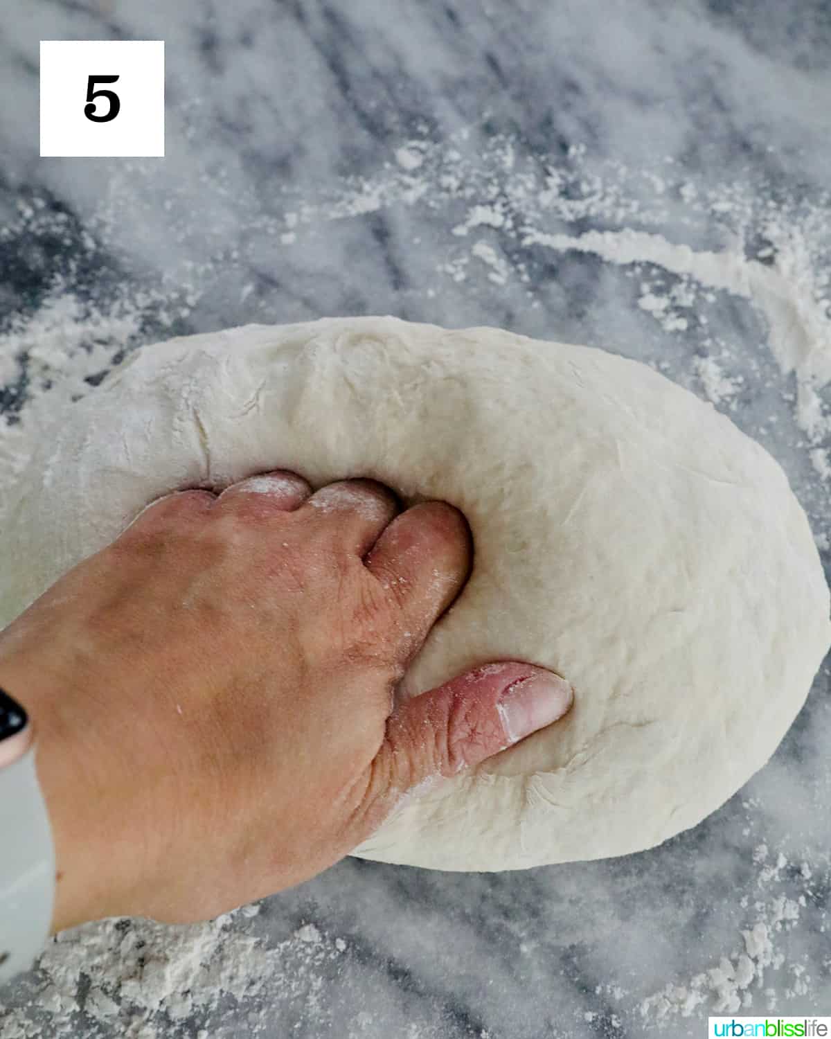A hand kneading homemade French bread dough ball on a floured marble countertop.