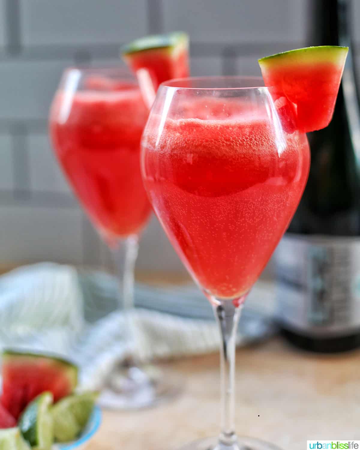 two glasses of watermelon mimosas with bottle of sparkling wine and watermelon slices in the background.