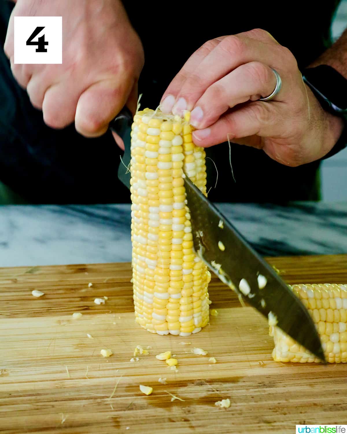 hands using a knife to slice down a corn on the cob on a cutting board.