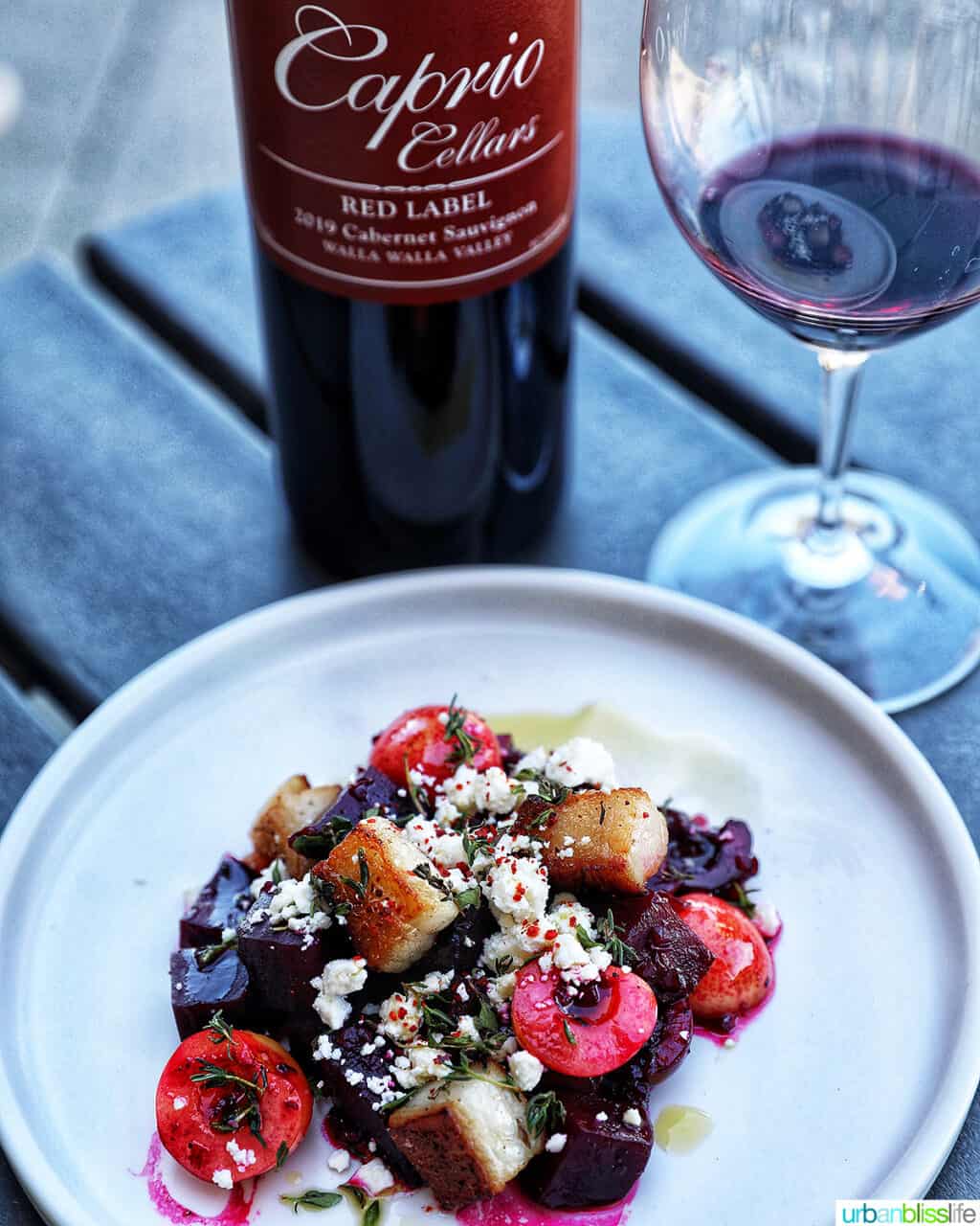 bottle and glass of red wine with a colorful beet, cherry, pork belly salad on a white plate.