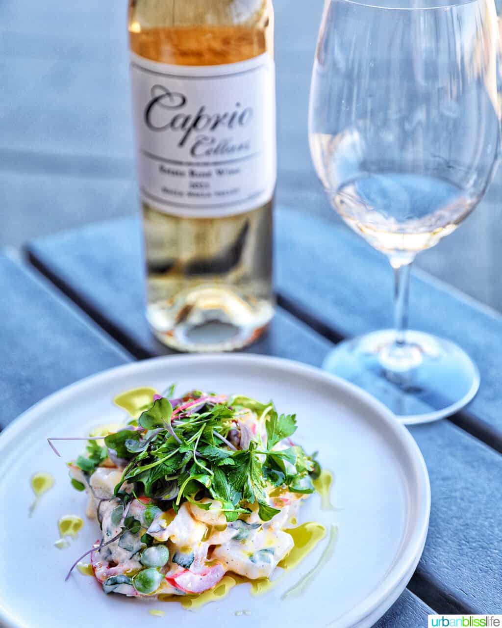 bottle of rosé wine, rosé wine in a glass, and a white plate topped with potato salad and microgreens.