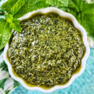 white scalloped bowl filled with mint pesto with mint leaves around the top of the bowl on a bright blue background.