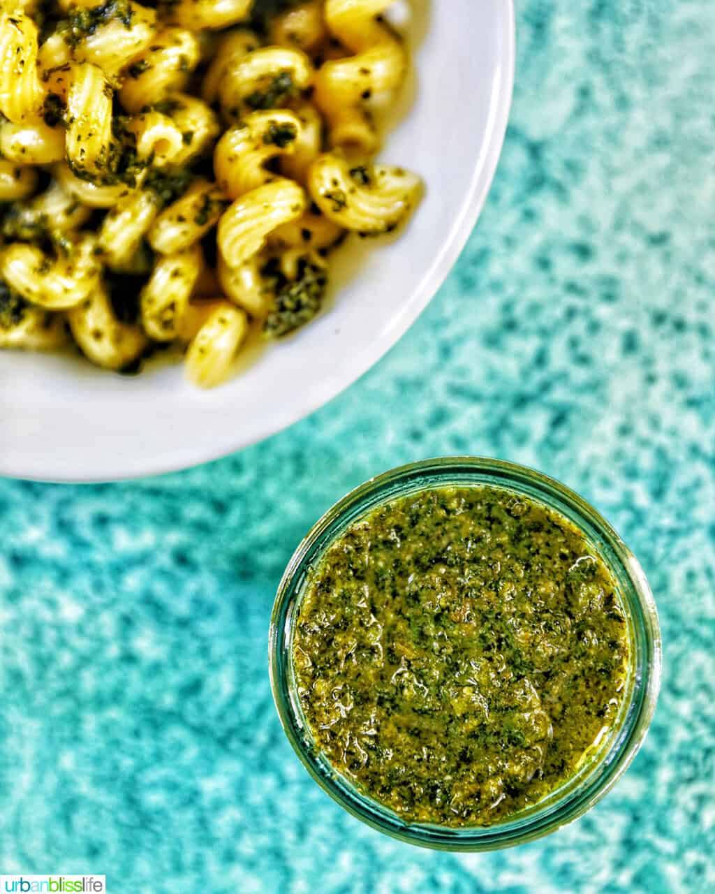 jar of mint pesto in the bottom right corner with bowl of pesto pasta in the top left corner, all on a bright blue background.