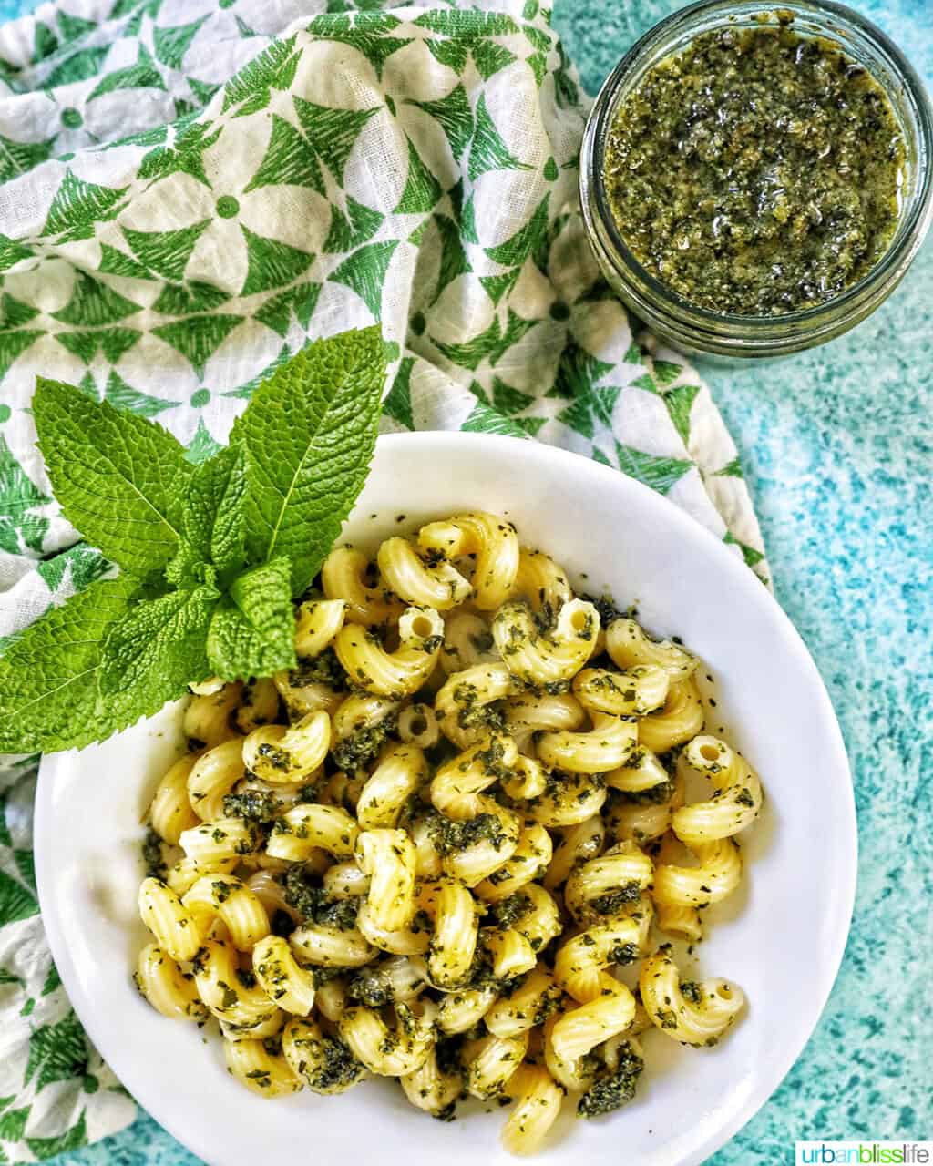 white bowl with pasta and mint pesto with mint leaves as garnish, with a jar of mint pesto in the top right corner, all on a bright blue background with green and white napkin to the left side.