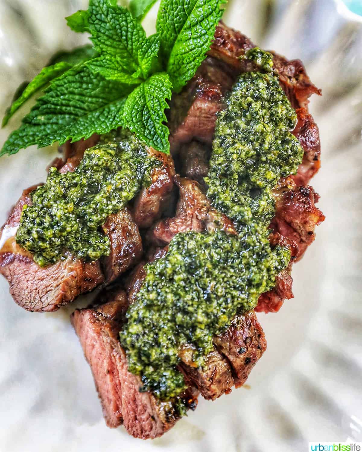 steak slices topped with mint pesto, with mint leaves as garnish on the top, on a white scalloped plate.