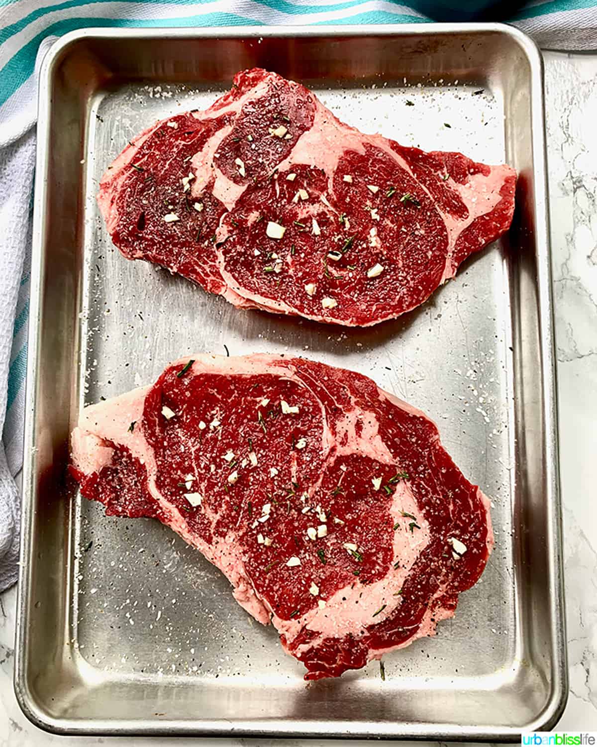 two rib eye steaks uncooked and seasoned on a metal dish.