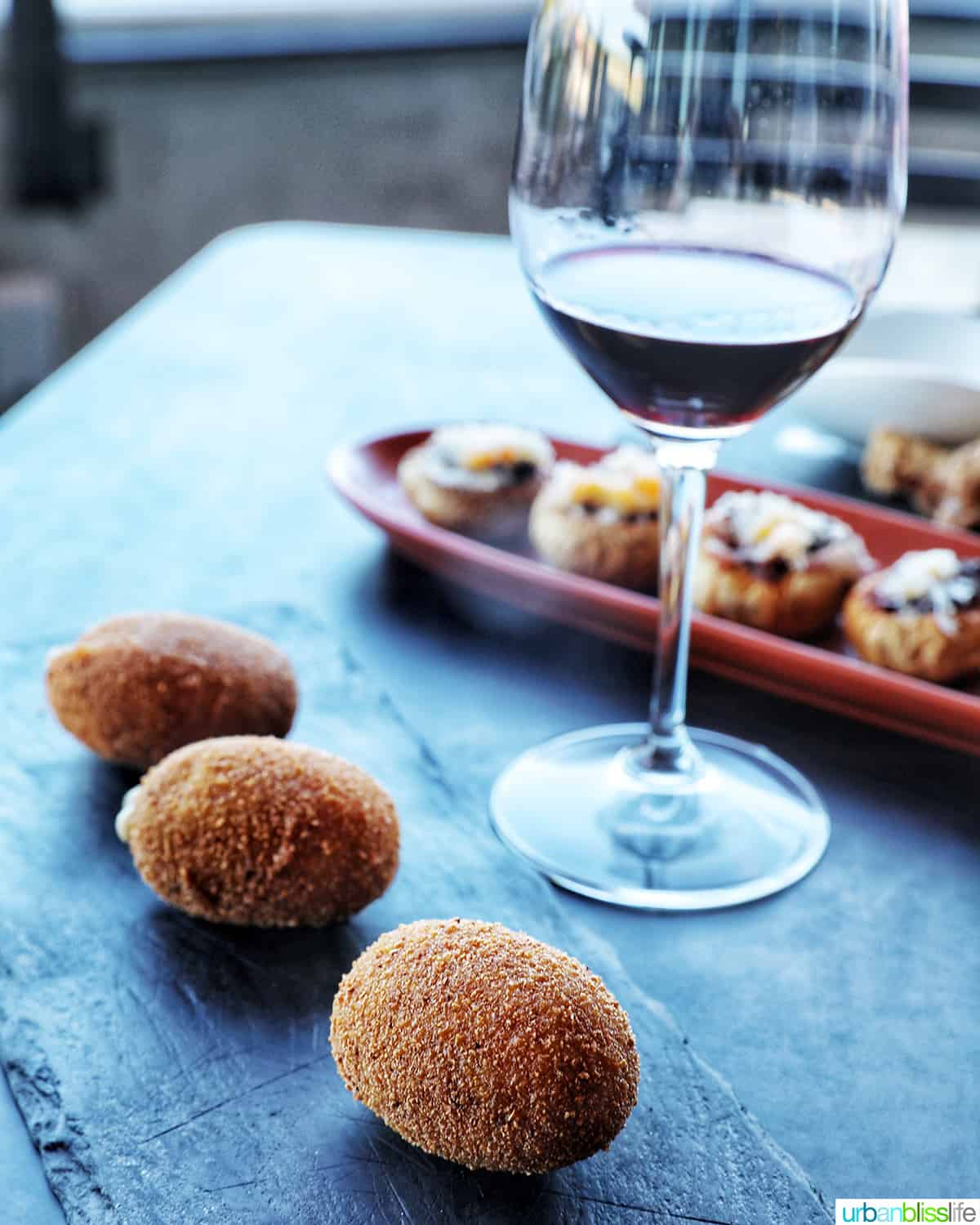three croquetas de jamon tapas on a black slate plate with glass of red wine and other tapas in the background.