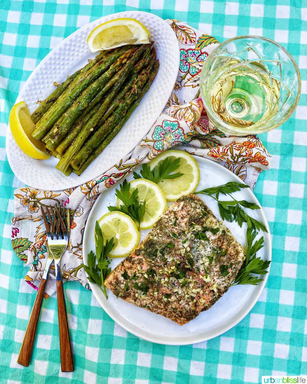 plate with grilled asparagus and lemon next to lemon slices and plate of grilled salmon with lemon and herbs, glass of white wine, forks, all on an aqua checkered tablecloth.