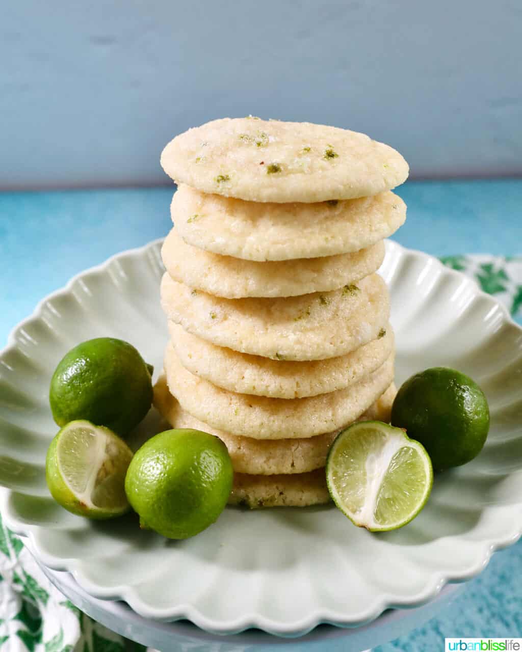 stack of key lime cookies on a scalloped plate with key limes around the cookies.