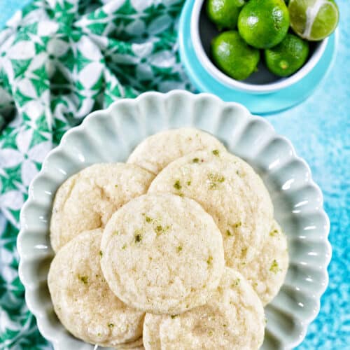 several key lime cookies on a scalloped plate with a bowl of key limes above and a bright blue background.