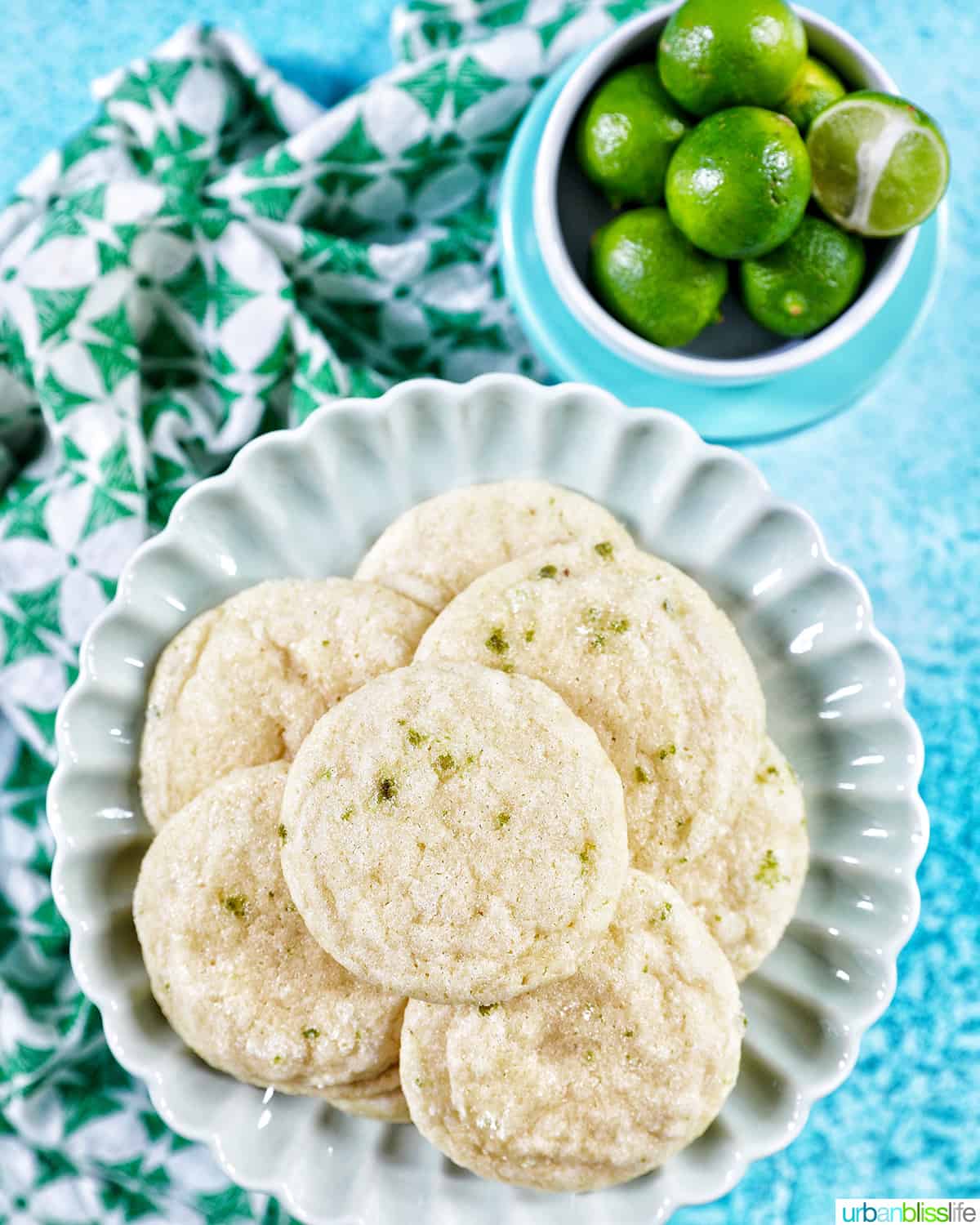 several key lime cookies on a scalloped plate with a bowl of key limes above and a bright blue background.