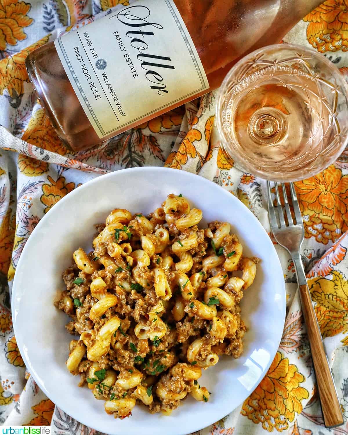 bowl of dairy free cheeseburger pasta on colorful napkins with bottle and glass of rosé wine.