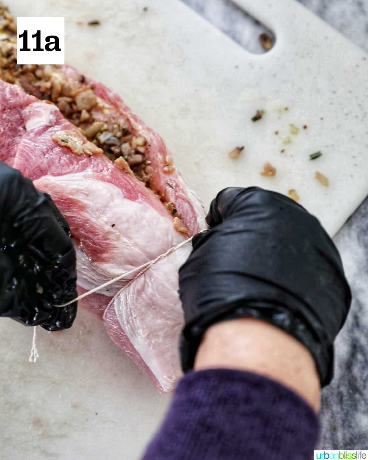 gloved hands tying kitchen twine around the stuffed and rolled pork loin on a white cutting board.